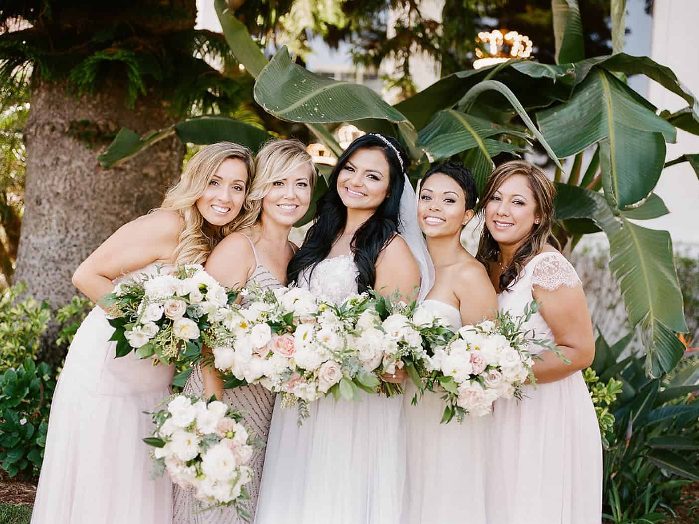 Bridesmaids and bride in elegant dresses holding floral bouquets at outdoor wedding celebration.