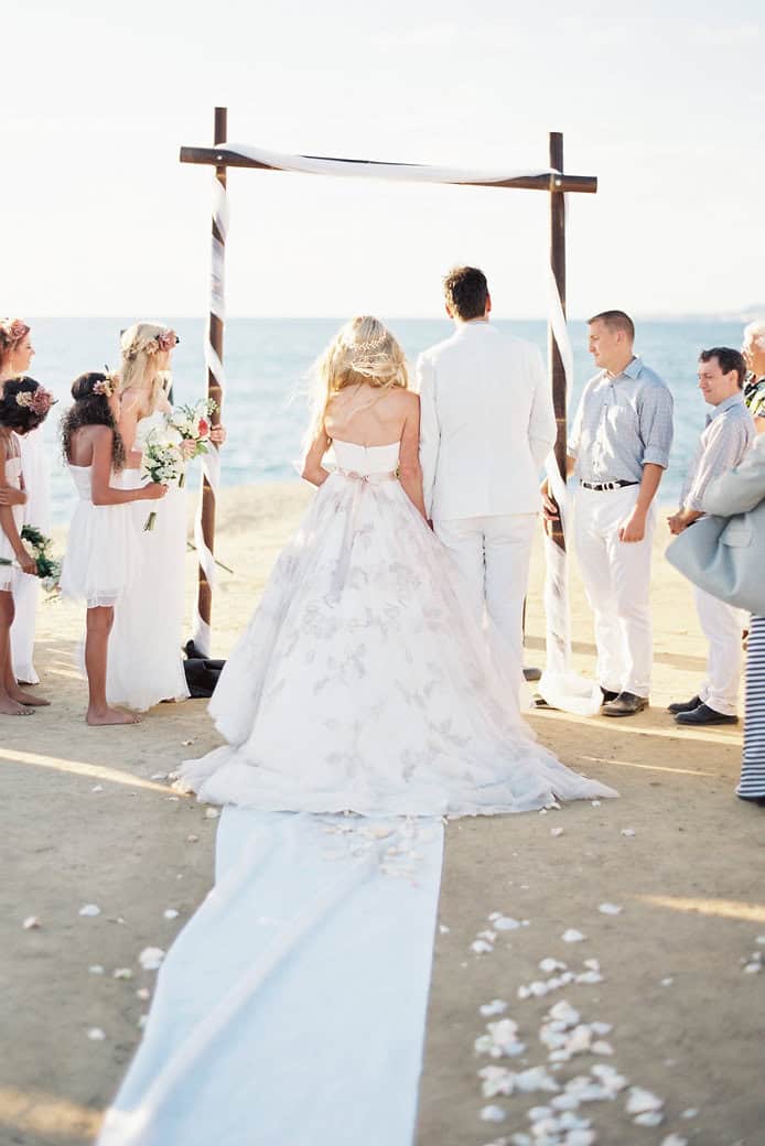 Bride and groom standing under a wooden arch on the beach during a wedding ceremony.